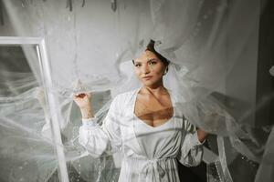 A beautiful brown-haired bride in a white dress poses for a photographer, standing under a veil in a white robe. Wedding photography, close-up portrait, chic hairstyle. photo