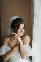 A brunette bride in a white dress holds her gorgeous curls and looks down. Open shoulders. Beautiful hands. Long veil. Morning of the bride. photo