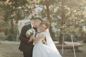 Wedding portrait. The groom in a black suit and the blonde bride are standing, hugging, posing holding a bouquet under a tree. Photo session in nature. Beautiful hair and makeup