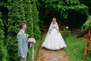 Portrait of the bride and groom standing against the background of green trees and holding a wedding bouquet of pink peonies. The bride goes to meet the groom. Stylish groom. Fashion and style. photo