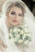 Rizado rubia novia en un blanco vestido, cubierto con un velo, poses para el cámara con un ramo de flores de rosas. retrato de el novia. hermosa maquillaje y cabello. Boda en naturaleza foto