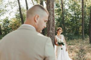 Wedding photo in nature. The bride is standing near a tree in a beautiful dress, holding a bouquet of white roses, looking at the camera. The bridegroom is in the foreground