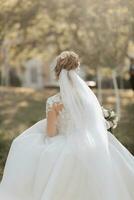 Wedding portrait. A blonde bride in a white dress with a train is walking, smiling and holding a bouquet and her wedding dress. Photo session in nature. Sun rays in the photo