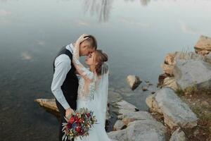 brides on the bank of the river, with bare legs hugging and kissing on a rock holding a bouquet of various flowers, red roses and blue flowers and a long white dress photo