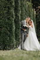 portrait of the bride and groom in nature. The groom is holding a bouquet, leaning against the bride, who is hugging him from behind, against the background of conifers. Stylish groom photo