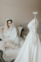 The blonde bride is dressed in a satin robe of seats and a sofa under a veil. Beautiful hair and make-up, open bust. Wedding portrait. A beautiful bride in a dressing gown poses next to her dress photo