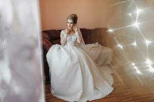 Blonde bride in long-sleeved lace dress sitting on leather sofa in her room, posing and wearing earrings. Beautiful hair and makeup, open shoulders. Wedding portrait. French manicure photo