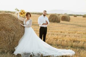 Wedding portrait of the bride and groom. The groom, tearing his shirt, stands behind the bride, near a bale of hay. Red-haired bride in a long dress with a bouquet of sunflowers. Stylish groom. Summer photo