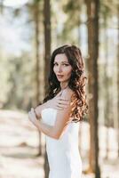 A girl with long black curly hair blowing in the wind is standing in a white silk dress. Standing in the spring forest against the background of large trees photo