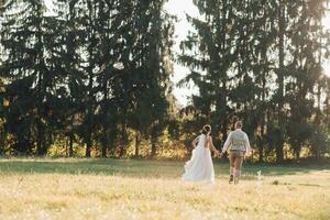 Happy bride and groom running in the field, holding hands, with their shoulders turned to the camera, big green Christmas trees in front of them. Beautiful light. Stylish groom. Pretty Girl photo