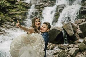 Portrait of a happy bride and groom near a waterfall. The groom holds the bride in his arms. Wedding photo session in nature. Photo session in the forest of the bride and groom.