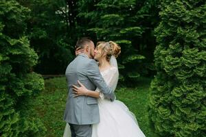 retrato de el novia y novio en pie en contra el antecedentes de verde árboles, abrazando y besando elegante novio. Moda y estilo. hermosa novia foto