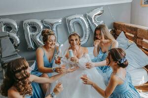 joven damas de honor en azul seda vestidos bebida champán en el de la novia habitación. hermosa mujer celebrando despedida de soltera fiesta sentado en cama y con champán. foto