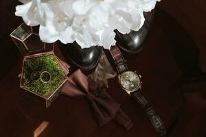 Morning of the groom and details, details of the groom on a dark background. men's watch, wedding rings in a glass box and wooden moss, a men's butterfly and a bouquet of white flowers photo