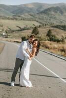 A stylish groom in a white shirt and a cute brunette bride in a white dress are hugging and looking at the camera on an asphalt road against the background of a forest and mountains. photo