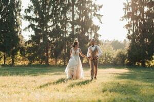 Happy newlyweds are running in the field, holding hands, looking at each other, behind them are big green Christmas trees. Beautiful light. Stylish groom. Pretty Girl photo
