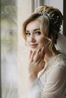 Blonde bride with elegant hairdo and tiara, wearing a robe, posing, looking at the camera, touching her face. Beautiful makeup. Elegant hairstyle. French manicure photo
