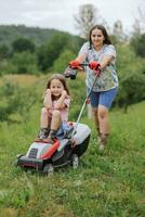 un mujer en botas con su niño en el formar de un juego corta el césped con un cortacésped en el jardín en contra el antecedentes de montañas y niebla, jardín herramientas concepto foto