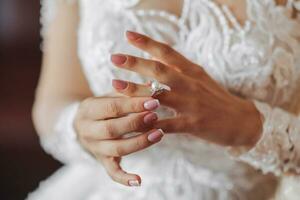 Blonde bride in long sleeve lace dress standing in her room, posing and putting on her engagement ring. Beautiful hair and makeup, open shoulders. Wedding portrait. French manicure photo