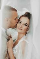 Wedding portrait of the bride and groom. Happy bride and groom gently hug each other under the veil, pose and kiss. Stylish groom. Beautiful young bride photo