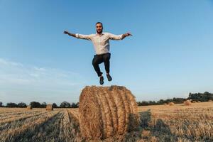 Wedding photo. Stylish groom in white shirt jumping down from hay bale and looking at camera. Bearded man. Style. Emotions photo