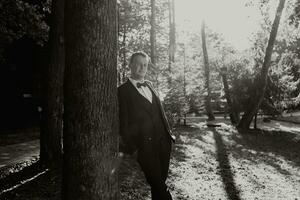 portrait of the groom in the forest in a wide shot, the groom leans his shoulder against a tree, the photo is backlit. black and white photo