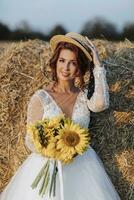 Wedding portrait. Red-haired bride in a white dress and hat, holding a bouquet of sunflowers, smiling and looking at the camera. Beautiful curls. Sincere smile. Elegant dress. photo