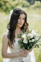 A beautiful bride with long curly hair in a chic dress, smiling, holding her wedding bouquet, looking into the lens. Portrait of the bride. Spring wedding. Natural makeup photo