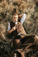 portrait of a stylish groom on a background of dry autumn grass. the concept of a rural wedding in the mountains, happy bohemian newlyweds. man relaxing lying on the grass photo