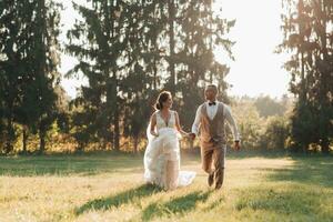 Happy newlyweds are running in the field, holding hands, looking at each other, behind them are big green Christmas trees. Beautiful light. Stylish groom. Pretty Girl photo