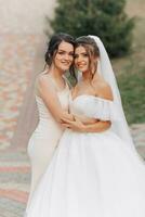 Wedding photo in nature. A brunette bride in a white long dress and her friend in a nude dress are hugging and smiling and posing. Young women. emotions