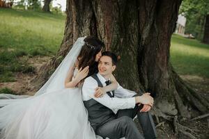 un hermosa novia con largo Rizado pelo en un elegante vestir abrazos el acicalar, sonrisas, mirando dentro el lente debajo un grande árbol. retrato de el novia y novio. primavera boda. natural maquillaje foto