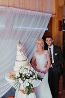 A white wedding cake, decorated with flowers and gold, stands on a glass stand. Fresh flowers for a wedding. sweets. The bride and groom near the cake photo