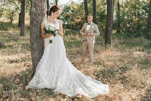 Boda foto en naturaleza. el novia es en pie cerca un árbol en un hermosa vestido, participación un ramo de flores en su manos, el novio es caminando detrás el novia. reunión en el bosque.