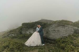 Happy wedding couple in the mountains near a big stone. The groom kisses the bride's hand. Wedding photo session in nature. Photo session in the forest of the bride and groom.