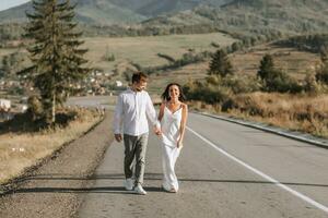 A stylish groom in a white shirt and a cute brunette bride in a white dress are walking on an asphalt road against the background of a forest and mountains. Wedding portrait of newlyweds. photo