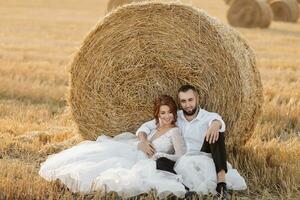 Wedding portrait. The bride and groom are sitting in an embrace on the ground, near a bale of hay. Red-haired bride in a long dress. Stylish groom. Summer. A sincere smile. On the background of hay photo
