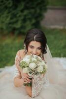 retrato de el novia en naturaleza. un morena novia en un blanco cordón vestir se sienta en el césped, posa, participación un ramo. hermosa pelo y constituir. abierto espalda. Boda disparo foto