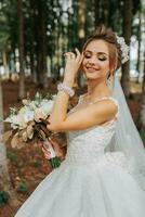 A beautiful young woman in a wedding dress between tall trees in the forest with a royal hairstyle and a chic tiara with a bouquet of flowers in her hands, a wedding in golden color photo
