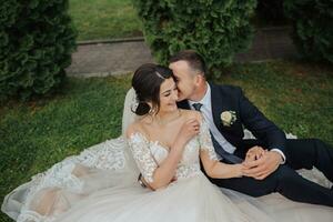 Portrait of the bride in nature. A brunette bride in a white lace dress and a stylish groom in a black suit are sitting on the grass, posing, holding a bouquet. Beautiful makeup. Open shoulders. photo