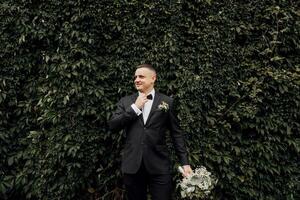 A happy groom in a black suit with a bouquet of flowers in his hand adjusts his tie on the background of a wall covered with green leaves photo