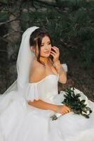 portrait of the bride in nature. A brunette bride in a white voluminous dress is sitting, posing near a coniferous tree, holding a bouquet of white roses. Beautiful hair and makeup. Warm light photo