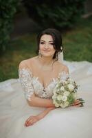 Portrait of the bride in nature. A brunette bride in a white lace dress sits on the grass, poses, holding a bouquet. Beautiful hair and makeup. Open shoulders. Wedding shooting photo