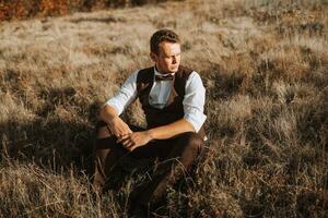 portrait of a stylish groom on a background of dry autumn grass. the concept of a rural wedding in the mountains, happy bohemian newlyweds. man relaxing sitting on the grass photo