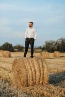 Wedding portrait photo. A stylish groom in a white shirt poses on a hay bale with his hands in his pockets. Bearded man. Style. photo