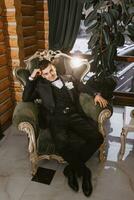 portrait of positive young groom in black suit with white shirt smiling and sitting on comfortable dark green sofa in modern hotel room on wedding day photo