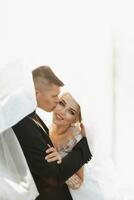 Wedding portrait. The groom in a black suit and the blonde bride are hugging, wrapped in the bride's veil. Long dress in the air. Photo session in nature. Beautiful hair and makeup