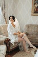 The brunette bride is sitting on the sofa in her room, a beautiful crown, putting on her wedding band, posing. Wedding portrait wedding photo. Beautiful makeup and hair. Morning of the bride photo