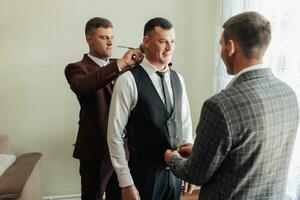 Photo of three men in classic suits. A handsome young man mends the shirt of another man, standing behind him. Business style. Stylish men