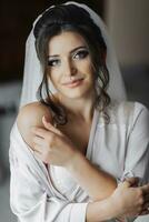 Brunette bride in a robe posing for the camera, looking into the camera. Open shoulders. Elegant hairstyle. Nice makeup. Voluminous veil. Morning of the bride. Beautiful hands of the bride photo